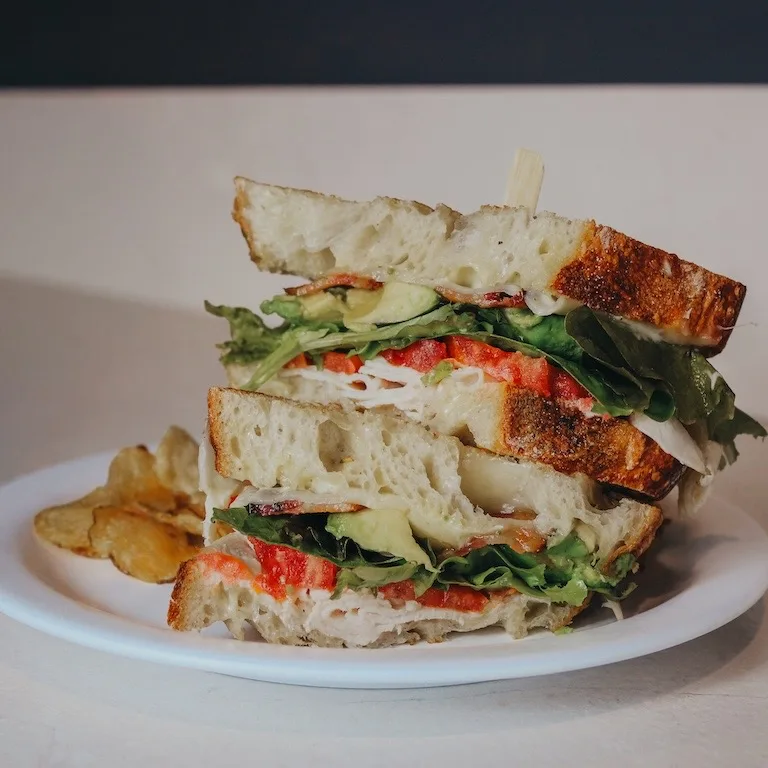 Stacked gourmet sandwich on artisan bread with chips at Niko’s Bakery & Cafe